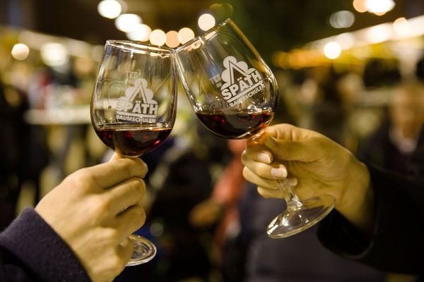 Zwei Hände stoßen mit Weingläsern an, die mit rotem Wein gefüllt sind und das Logo "SPÄTH-Baumschulen" tragen. Im unscharfen Hintergrund sind warme Lichter und eine Restaurantumgebung zu erkennen.