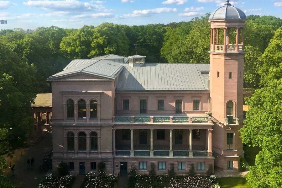 Schloss Biesdorf mit seiner charakteristischen rötlichen Backsteinfassade, einem markanten Turm mit Kuppeldach und einem Balkon mit Säulen, umgeben von grünen Bäumen unter blauem Himmel mit weißen Wolken.