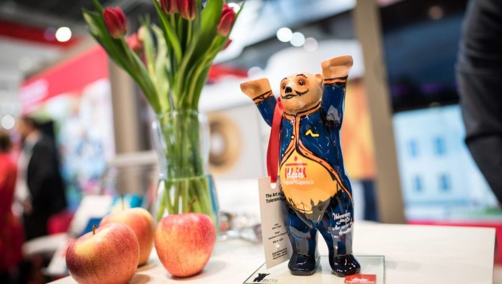 Eine kleine Buddybär-Figur in blau-gelber Bemalung steht auf einem weißen Tisch neben roten Tulpen in einer Glasvase und zwei Äpfeln. Im Hintergrund ist unscharf ein Ausstellungsraum zu erkennen.