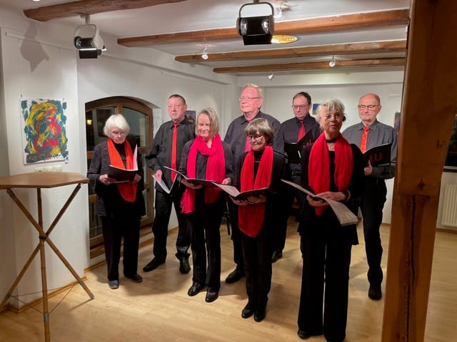 Acht Personen in schwarzer Kleidung mit roten Schals oder Stolen stehen in einer Reihe und halten Notenblätter in den Händen, während sie in einem Raum mit Holzbalken an der Decke singen oder proben.