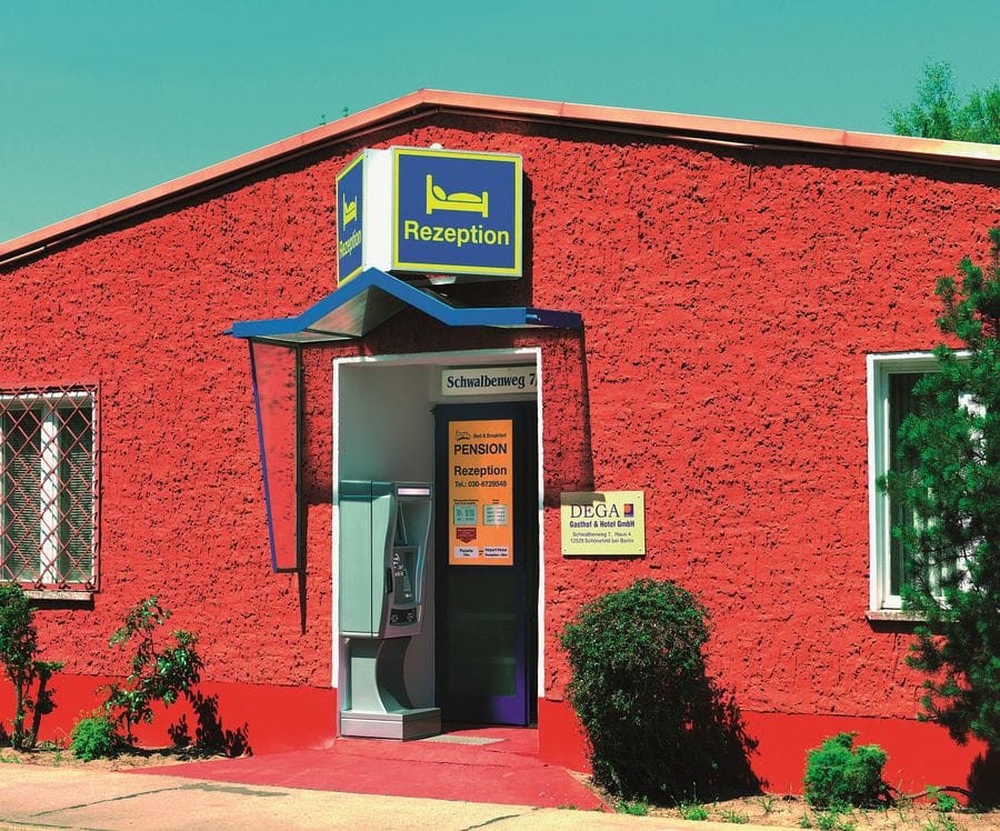Eingang einer Pension mit roter Fassade am Schwalbenweg 7, mit blau-gelber Rezeption-Beschilderung über der Eingangstür und einem Telefonhäuschen davor. Das einstöckige Gebäude hat ein rotes Ziegeldach und ist von Grünpflanzen umgeben.