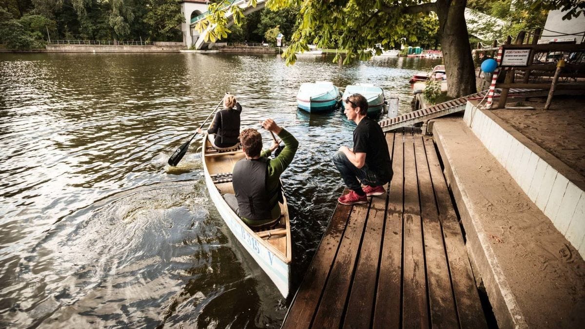 Zwei Personen steigen von einem Holzsteg in ein weißes Kanu auf der Spree in Berlin Treptow nahe der Elsenbrücke ein. Im Hintergrund sind weitere Boote und ein Bootsverleih unter schattigen Bäumen am Ufer zu sehen.