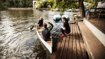 Zwei Personen steigen von einem Holzsteg in ein weißes Kanu auf der Spree in Berlin Treptow nahe der Elsenbrücke ein. Im Hintergrund sind weitere Boote und ein Bootsverleih unter schattigen Bäumen am Ufer zu sehen.