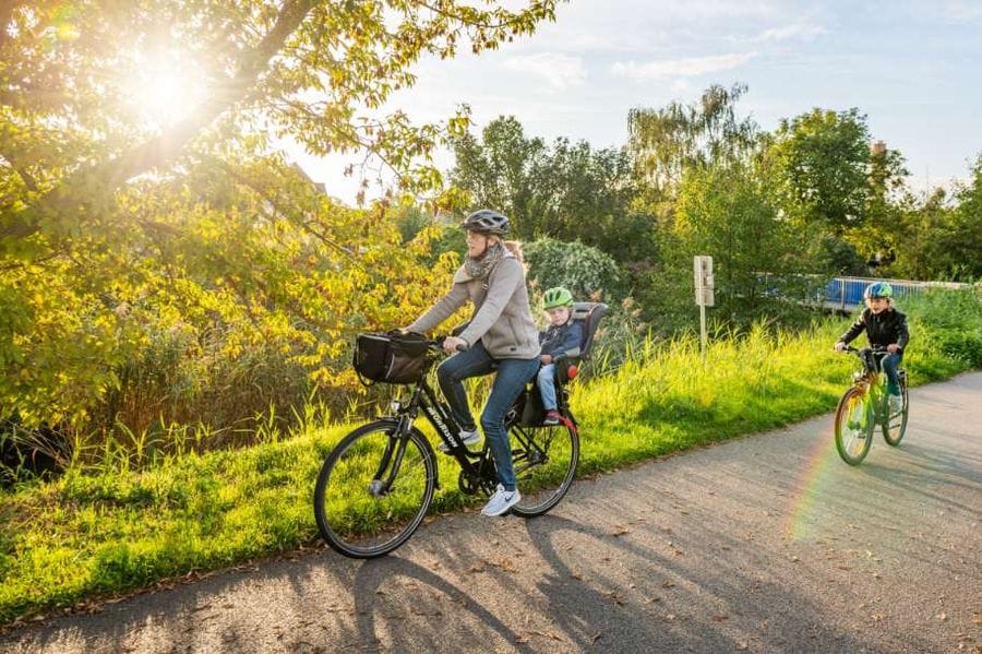 Eine Frau fährt mit einem Fahrrad mit Kindersitz, in dem ein Kind mit grünem Helm sitzt, auf einem asphaltierten Weg durch eine grüne Landschaft. Ein weiteres Kind fährt rechts auf einem eigenen Fahrrad neben ihnen, während die Sonne durch die Bäume scheint.