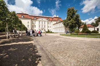 Schloss Köpenick mit seiner weißen Barockfassade und dem roten Ziegeldach, davor ein gepflasterter Innenhof mit einer Gruppe von Touristen und parkähnlichen Grünanlagen mit Bäumen.