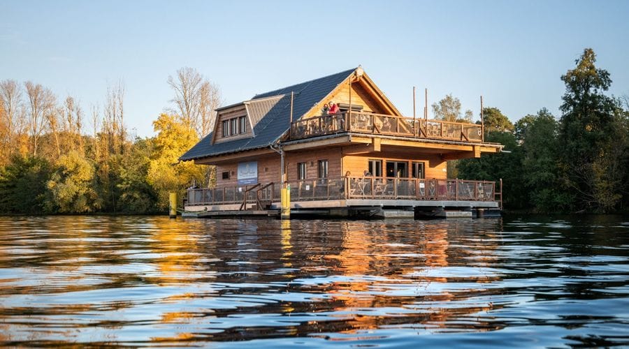 Schwimmendes Restaurant Spreearche auf dem Müggelsee mit mehrstöckiger Konstruktion, Balkonen und Terrassen, umgeben von herbstlich gefärbten Bäumen bei warmem Sonnenlicht.