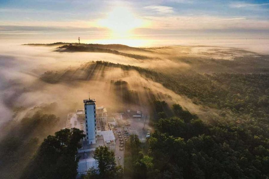 Luftaufnahme der Müggelberge bei Sonnenaufgang mit dichtem Nebel, der zwischen den bewaldeten Hügeln liegt und einen weißen Aussichtsturm sowie weitere Gebäude umhüllt. Im Hintergrund sind weitere Hügelketten und ein Funkturm durch den goldenen Morgennebel zu erkennen.