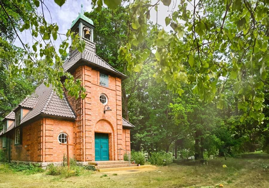 Die Waldkapelle Hessenwinkel steht inmitten grüner Bäume und zeigt sich als backsteinerner Kirchenbau mit einem hohen Turm, grünem Kupferdach und türkisfarbener Eingangstür. Das Gebäude ist von einer parkähnlichen Wiese und dichtem Laubwald umgeben.