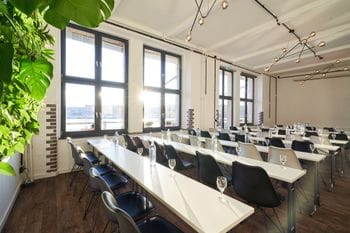 Tagungsraum mit mehreren Tischreihen und Stühlen. Auf den Tischen stehen Gläser. Die Sonne scheint durch die Fenster.