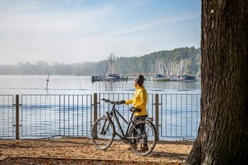 Eine Person in gelber Jacke steht mit ihrem Fahrrad an der Uferpromenade in Köpenick und blickt auf die Spree mit Segelbooten und bewaldeten Ufern. Im Vordergrund ist ein großer Baumstamm zu sehen, dahinter erstreckt sich das Geländer der Promenade mit Blick auf den Yachthafen.