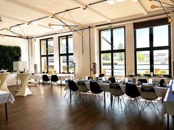 Moderner Veranstaltungsraum mit dunklem Holzboden, schwarzen Stühlen an weißen Tischen, großen Fenstern und einer Lichterkette an der Decke. Links im Raum befinden sich weiße Stehtische und eine grüne Pflanzenwand.