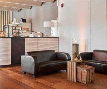 Moderner Wohnbereich mit zwei schwarzen Ledersofas, einem rustikalen Holzwürfel als Beistelltisch und einer offenen Küche mit hellen Holzfronten im Hintergrund. Der Raum hat Holzbalkendecke, Parkettboden und warme Beleuchtung.