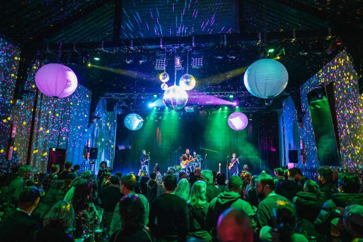 Eine Musikband bestehend aus drei Personen spielt auf einer Bühne vor einem Publikum in einem Club mit bunter Beleuchtung und hängenden Lampions. Die Bühne ist in grünes und blaues Licht getaucht, während das Publikum im Vordergrund der Aufführung zuschaut.