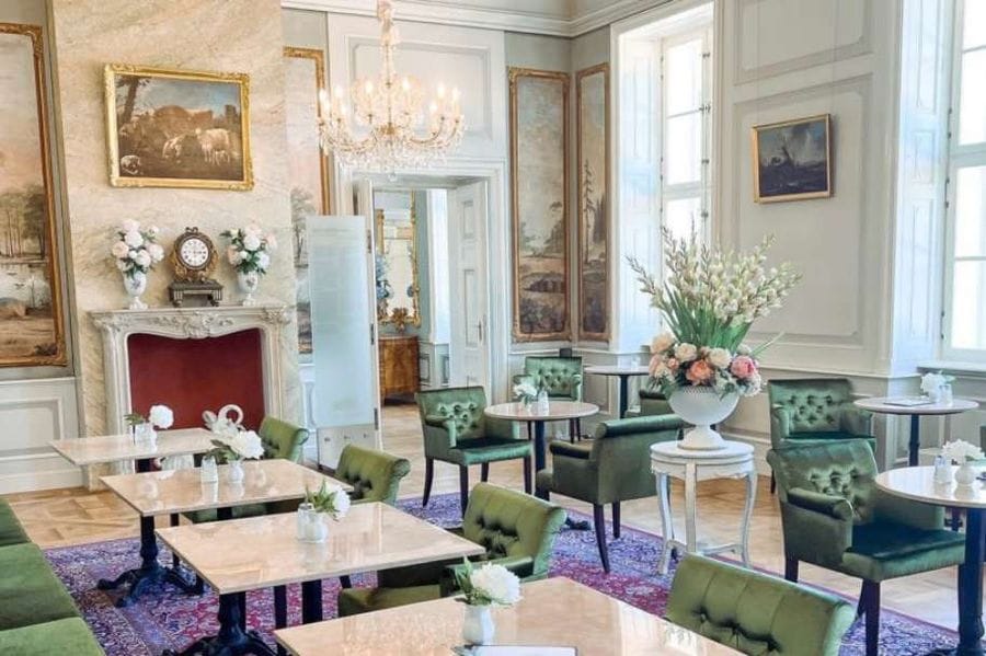 Eleganter Innenraum des Café Schloss Friedrichsfelde mit grünen Polstersesseln, hellen Marmortischen und klassischer Einrichtung. Der Raum verfügt über einen Kamin, Kronleuchter, historische Gemälde an den Wänden und große Fenster, die natürliches Licht hereinlassen.