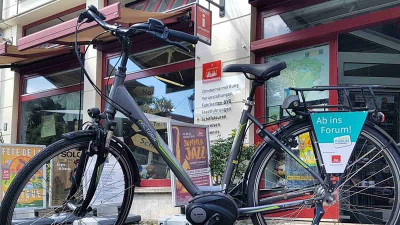 Zwei E-Bikes stehen vor der Touristeninformation Treptow-Köpenick mit großen Fenstern, in denen Informationsbroschüren und Plakate für lokale Veranstaltungen ausgestellt sind.