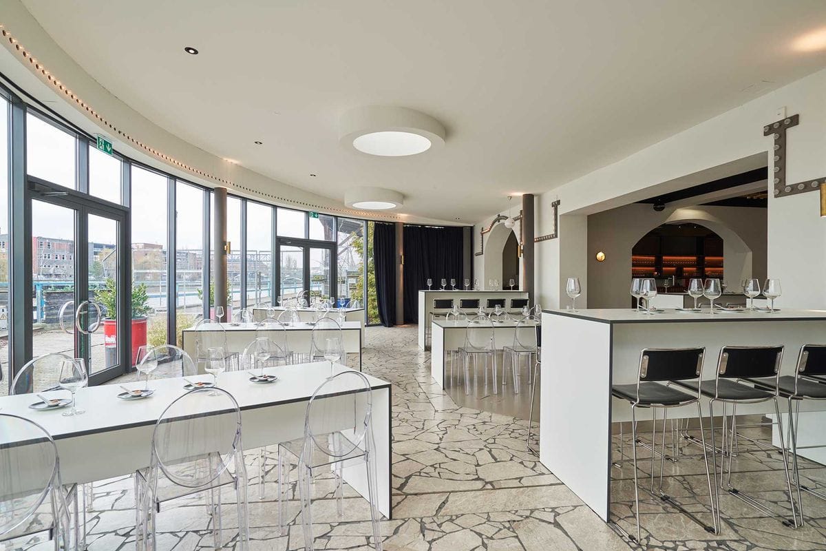 Moderner Restaurantinnenraum mit weißen Tischen, durchsichtigen Stühlen und einer weißen Theke mit schwarzen Barhockern. Der Raum verfügt über große Fenster zur Straße, helle Deckenbeleuchtung und einen gemusterten Steinboden.