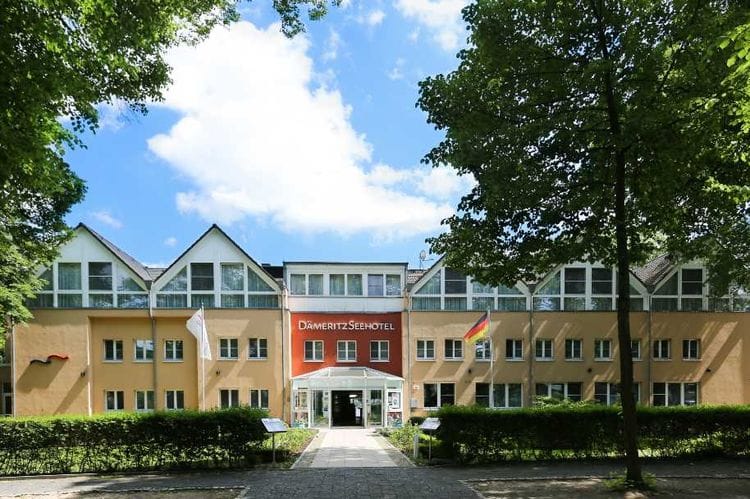 Das DämeritzSeehotel ist ein mehrstöckiges Gebäude mit beiger Fassade und markanter roter Eingangsfassade, umgeben von grünen Bäumen und gepflegten Grünanlagen. Vor dem Hoteleingang mit Glasvorbau führt ein gepflasterter Weg zum Gebäude.