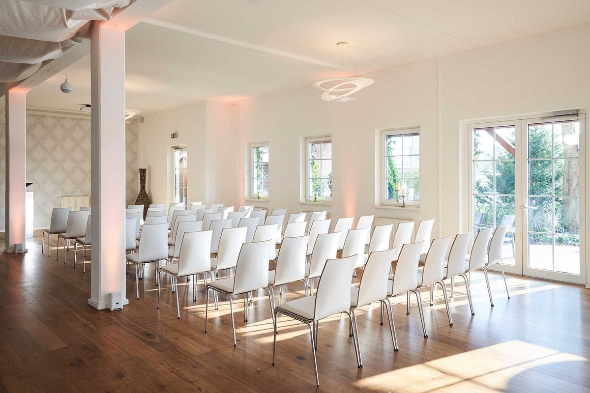 Ein heller Veranstaltungsraum mit etwa 30 weißen Stühlen in Reihen aufgestellt, weißen Säulen, dunklem Holzboden und großen Fenstern mit Blick ins Grüne. An der Decke hängt ein moderner Ring-Kronleuchter und rosa Lichtakzente beleuchten die weißen Wände.