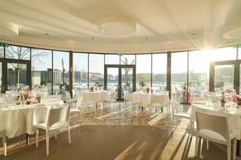 Das Restaurant "Lighthouse Café" mit runden Tischen, weißen Tischdecken und Stühlen, großen Fenstern mit Blick auf eine Hafenlandschaft und geometrisch gemustertem Boden. Das warme Tageslicht wirft Schatten auf den Boden.