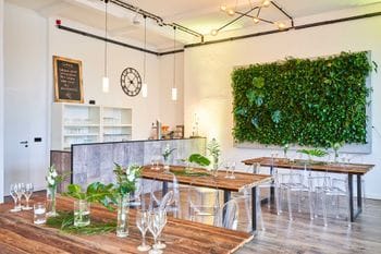 Modernes Restaurant mit Holztischen, transparenten Stühlen und einer großen grünen Pflanzenwand an der rechten Wand. Der Raum verfügt über eine offene Küche mit Betontheke, industrielle Deckenbeleuchtung und eine Schiefertafel mit Speisenangebot an der linken Wand.