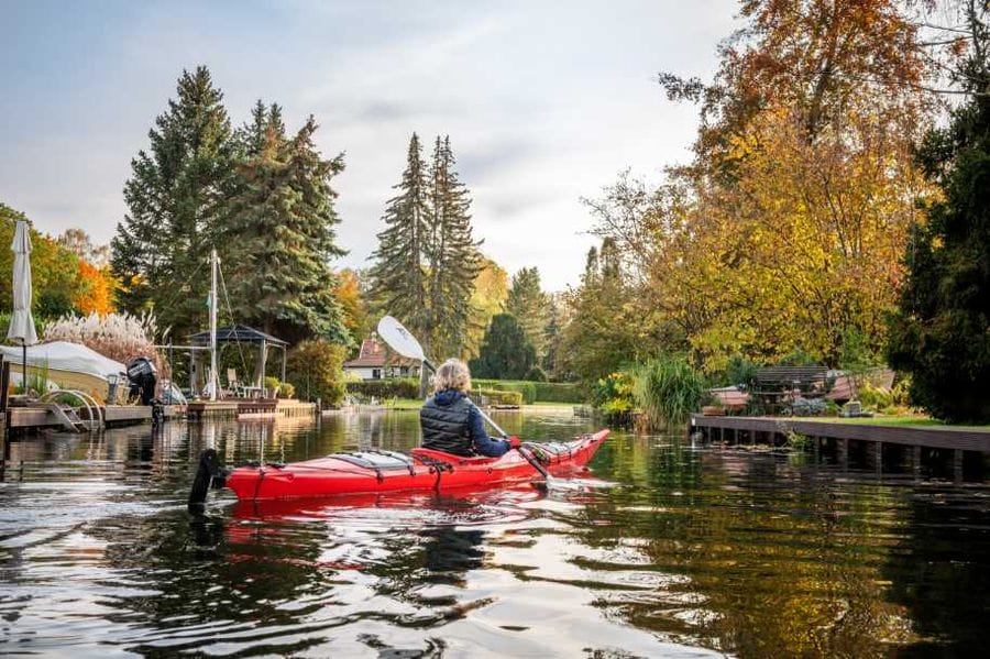 Eine Person paddelt in einem roten Kajak durch die ruhigen Kanäle von Neu Venedig, umgeben von herbstlich gefärbten Bäumen und traditionellen Bootsstegen. Im Hintergrund sind Boote, Häuser und die charakteristische Wasserlandschaft mit Holzstegen zu sehen.