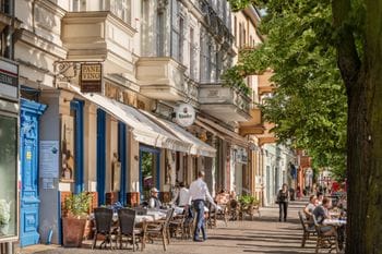 Die Bölschestraße mit historischen Gebäuden und Restaurants, vor denen Menschen an Tischen im Freien sitzen und speisen. Links ist ein Restaurant mit blauer Fassade namens "Pane Vino" zu sehen, rechts stehen große grüne Bäume entlang des Gehwegs.