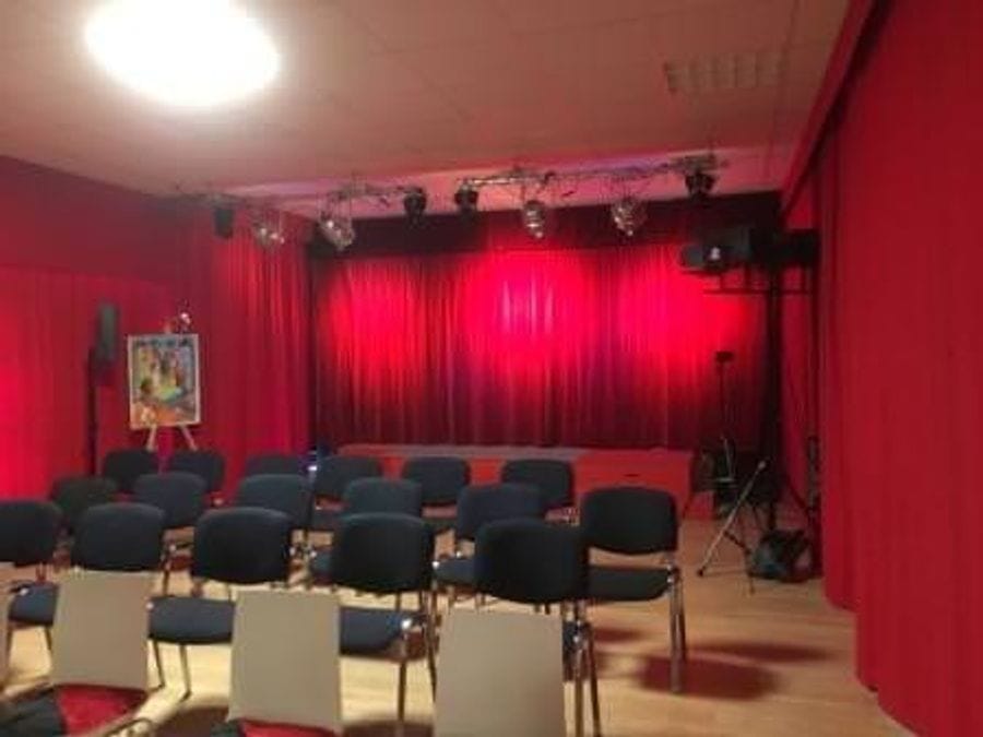 Innenansicht des Stadttheaters Köpenick mit schwarzen Stühlen im Zuschauerraum und einer kleinen Bühne mit rotem Vorhang und Beleuchtungsanlage.