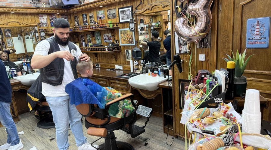Ein Friseur mit Bart schneidet in einem traditionellen Barbershop in der Altstadt Köpenick einem Kind die Haare. Der Shop ist mit dunklem Holz eingerichtet und verfügt über klassische Friseurstühle, Spiegel und zahlreiche Dekorationen an den Wänden.