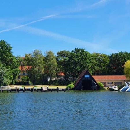 Blick über das Wasser auf das Waldhotel Am See mit roten Ziegeldächern zwischen grünen Bäumen, davor ein Holzsteg und weiße Motorboote am Ufer.