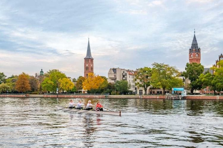 Blick auf die Altstadt Köpenick mit dem charakteristischen Rathaus und der Kirche im Hintergrund, während Ruderer in einem Boot auf der Dahme fahren. Die historischen Gebäude sind von herbstlichen Bäumen umgeben und spiegeln sich im Wasser.