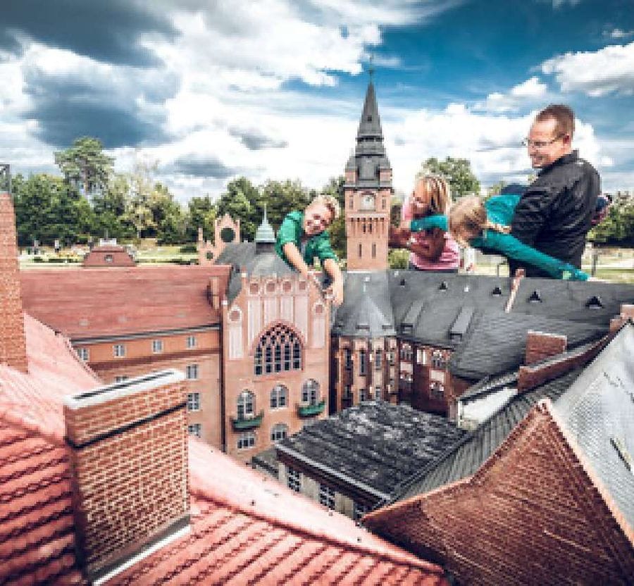 Eine Familie betrachtet und das Miniaturmodell vom Rathaus Köpenick im Freizeitpark Wuhlheide. Im Hintergrund sind unter blauem Himmel mit weißen Wolken Bäume und ein echter Kirchturm zu sehen.