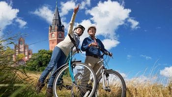 Zwei Fahrradfahrer mit Helmen stehen mit ihren Fahrrädern auf einer Wiese und einer zeigt mit dem Finger nach oben, während im Hintergrund ein historischer Rathausturm mit roter Backsteinfassade und Uhr zu sehen ist.