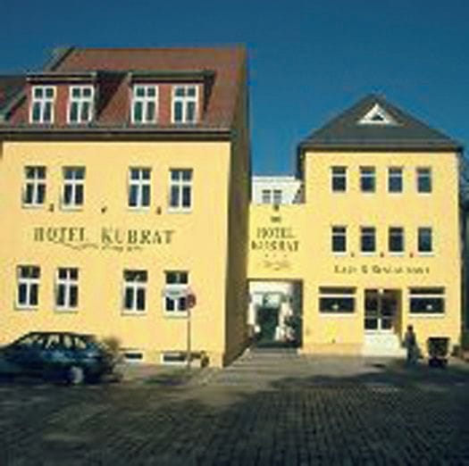 Zwei gelbe Gebäude des Hotel Kubrat in der Altstadt Köpenick bei blauem Himmel, mit gepflastertem Vorplatz und einem geparkten Auto vor dem Eingang.