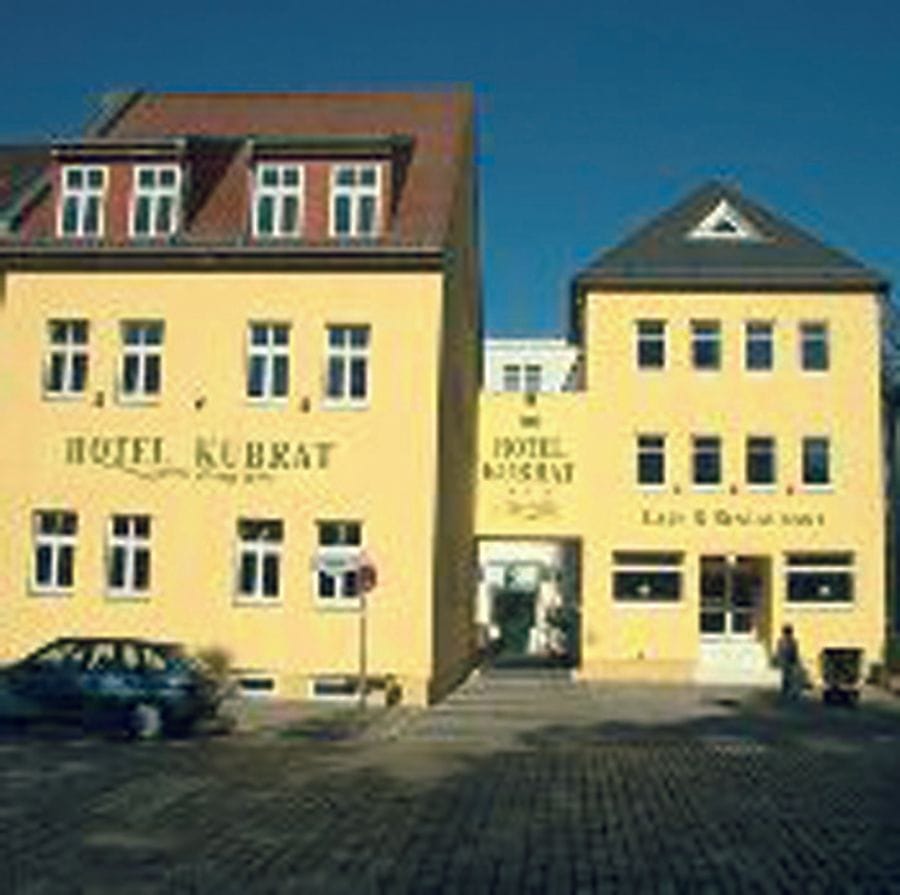 Zwei gelbe Gebäude des Hotel Kubrat in der Altstadt Köpenick bei blauem Himmel, mit gepflastertem Vorplatz und einem geparkten Auto vor dem Eingang.