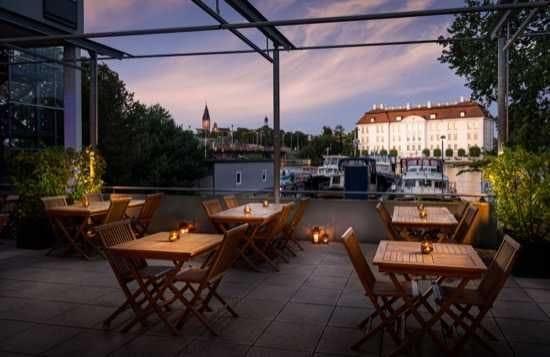 Terrasse des Hotel Berlin Köpenick by Leonardo Hotels mit Holztischen und -stühlen unter einem Glasdach, im Hintergrund sind die Spree mit Booten und historische Gebäude von Köpenick in der Abenddämmerung zu sehen.