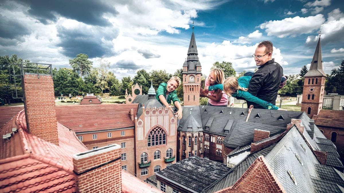 Eine Familie mit drei Kindern betrachtet ein Miniaturmodell des Rathaus von Köpenick in der Miniaturwelt Wuhlheide unter blauem Himmel mit weißen Wolken.