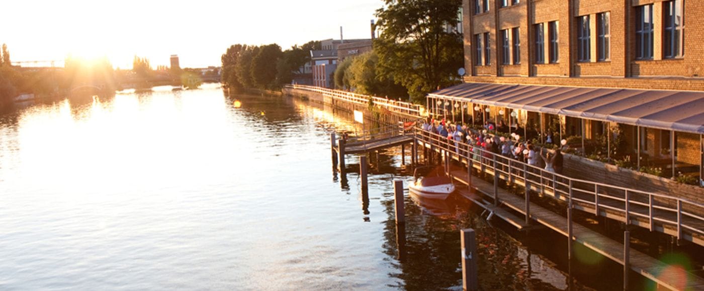 Sonnenuntergang an der Spree mit backsteinerner Uferpromenade in Schöneweide, auf der Menschen an Geländern stehen und auf das Wasser blicken. Im Vordergrund liegt ein kleines Boot am Steg, während sich die warme Abendsonne im Wasser spiegelt.