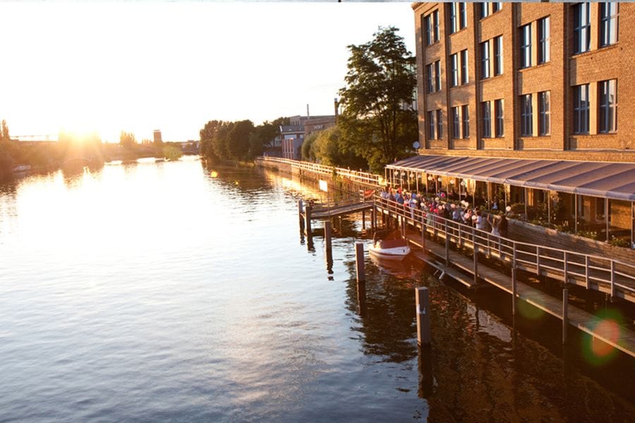 Sonnenuntergang an der Spree mit backsteinerner Uferpromenade in Schöneweide, auf der Menschen an Geländern stehen und auf das Wasser blicken. Im Vordergrund liegt ein kleines Boot am Steg, während sich die warme Abendsonne im Wasser spiegelt.