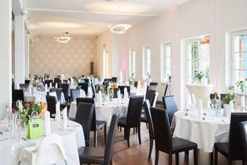 Ein elegant eingerichteter Restaurantsaal mit weißen Tischdecken, schwarzen Lederstühlen und kleinen Blumengestecken auf den Tischen. Der helle Raum verfügt über große Fenster, moderne Hängeleuchten und eine gemusterte Akzentwand.
