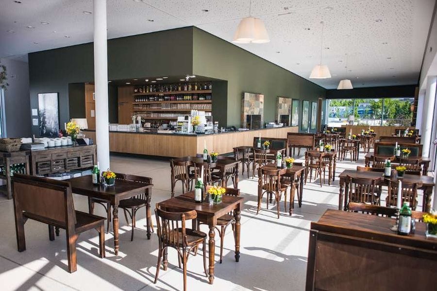 Innenansicht des Restaurant Giardino Verde in den Gärten der Welt mit brauen Holztischen und -stühlen im Gastraum. Im Hintergrund ist eine Bar mit grüner Wand und Getränkeregalen zu sehen, durch große Fenster fällt natürliches Licht in den modernen Restaurantbereich.