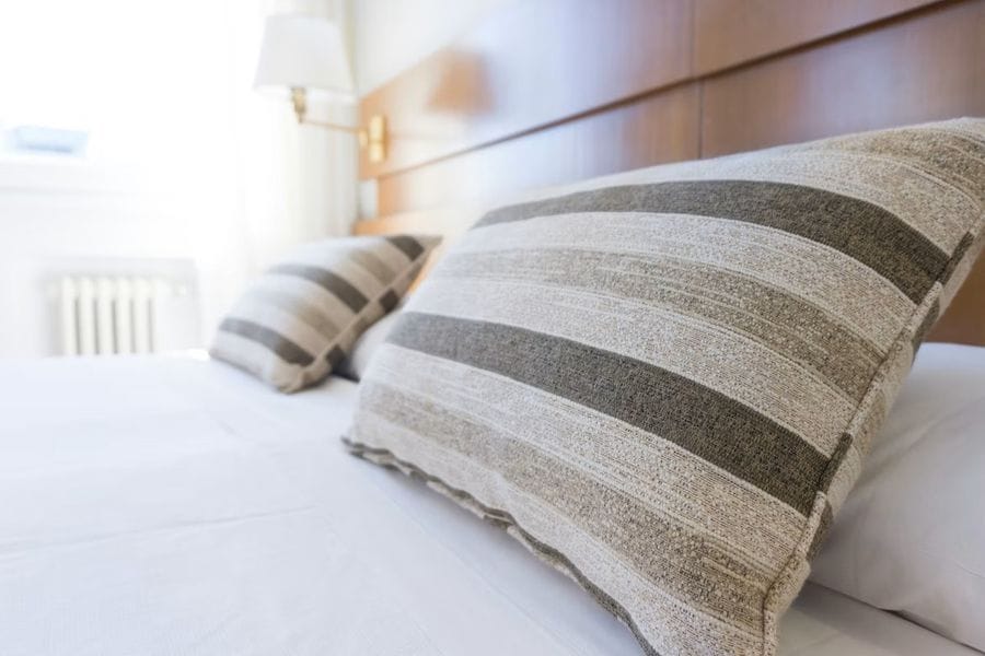 Zwei gestreifte Kissen in Grau- und Beigetönen liegen auf einem weißen Bett vor einem hölzernen Kopfteil. Im Hintergrund ist unscharf eine Lampe und helles Tageslicht durch ein Fenster zu erkennen.
