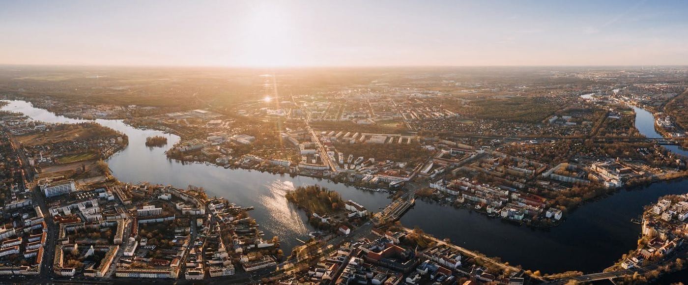 Luftaufnahme von Köpenick bei Sonnenuntergang, die den Stadtteil mit seinen Wasserwegen, der dichten Wohnbebauung und der charakteristischen Flusslandschaft zeigt. Die warme Abendbeleuchtung hebt die Struktur der Stadt und die sich schlängelnden Gewässer hervor.