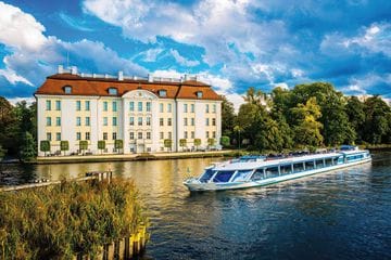 Ein weißes Ausflugsschiff der Reederei Stern und Kreis fährt auf dem Fluss Dahme vor dem barocken Schloss Köpenick mit seiner charakteristischen weißen Fassade und dem roten Ziegeldach. Das historische Schloss ist von grünen Bäumen umgeben und spiegelt sich im Wasser wider.
