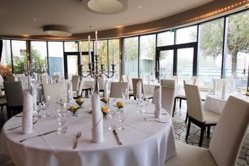 Elegant gedeckter Hochzeitssaal im Lighthouse Cafe Berlin mit weißen Tischdecken, cremefarbenen Stühlen und Kerzenleuchtern. Der lichtdurchflutete Raum verfügt über bodentiefe Fenster mit Blick ins Grüne und moderne Deckenbeleuchtung.