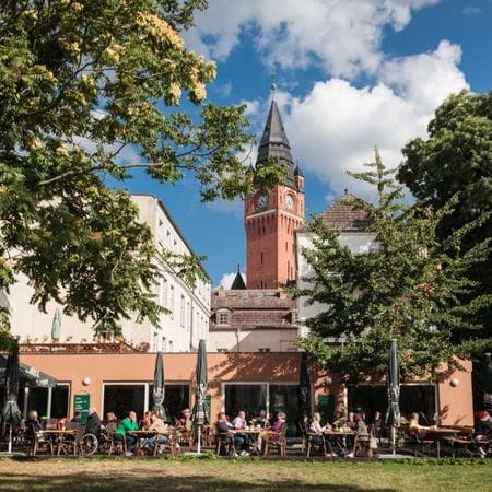Menschen sitzen an Tischen vor dem Café Luise im Freien, umgeben von grünen Bäumen, im Hintergrund der rote Backsteinturm vom Rathaus Köpenick. 