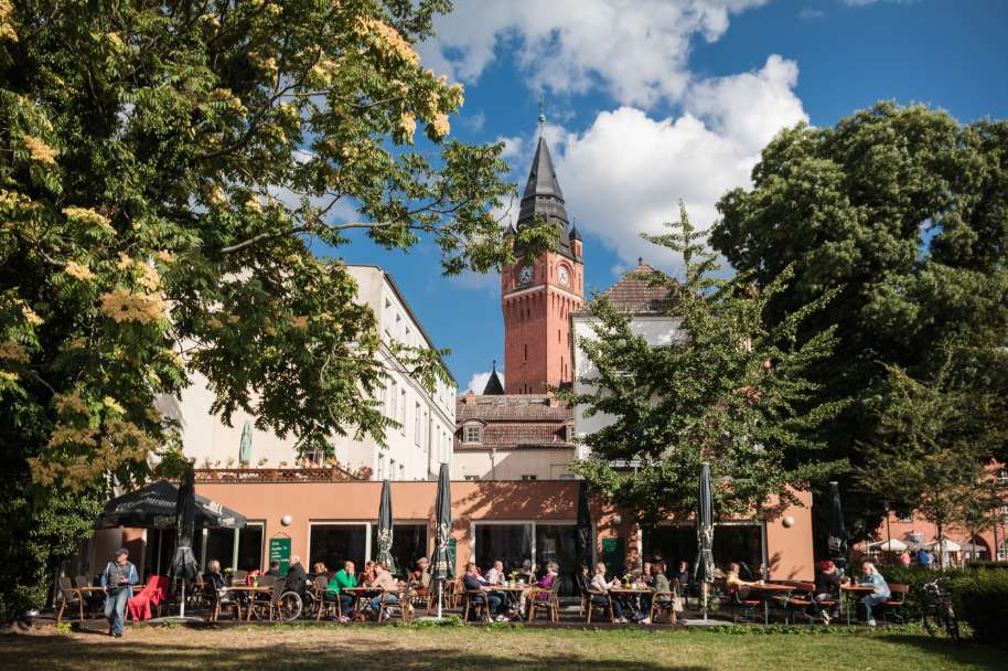 Menschen sitzen an Tischen vor dem Café Luise im Freien, umgeben von grünen Bäumen, im Hintergrund der rote Backsteinturm vom Rathaus Köpenick. 